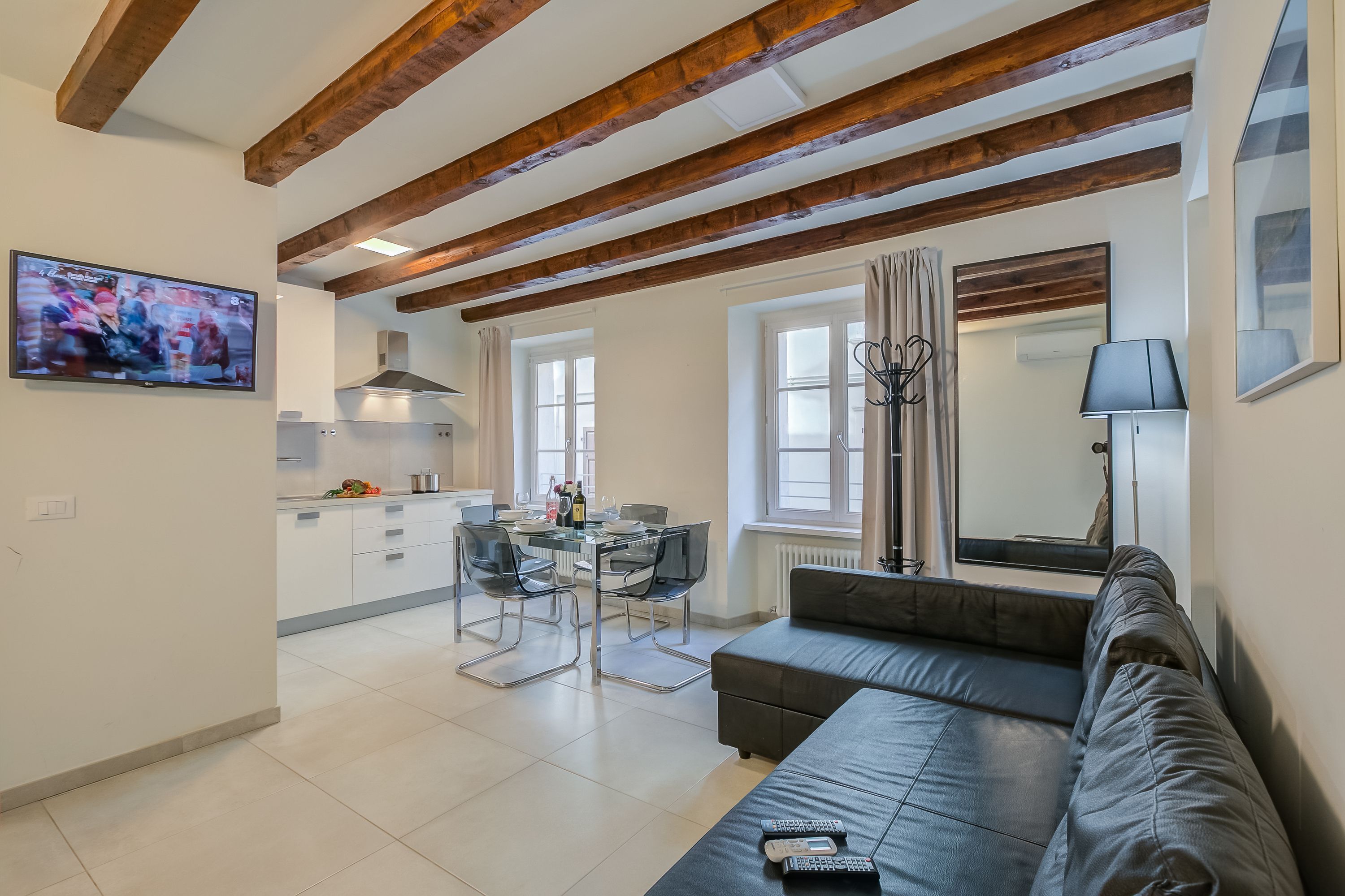 A modern living room with a sofa, a TV mounted on the wall, a dining area with a table and chairs, and a kitchen in the background.