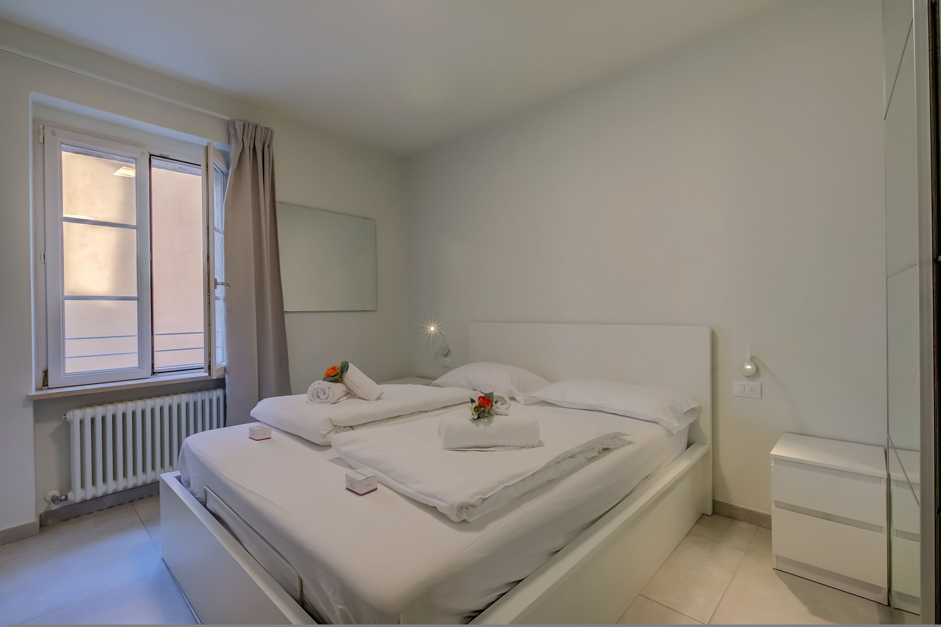 Una cama bien hecha con sábanas blancas, una mesita de noche con lámpara y una ventana con cortinas.
