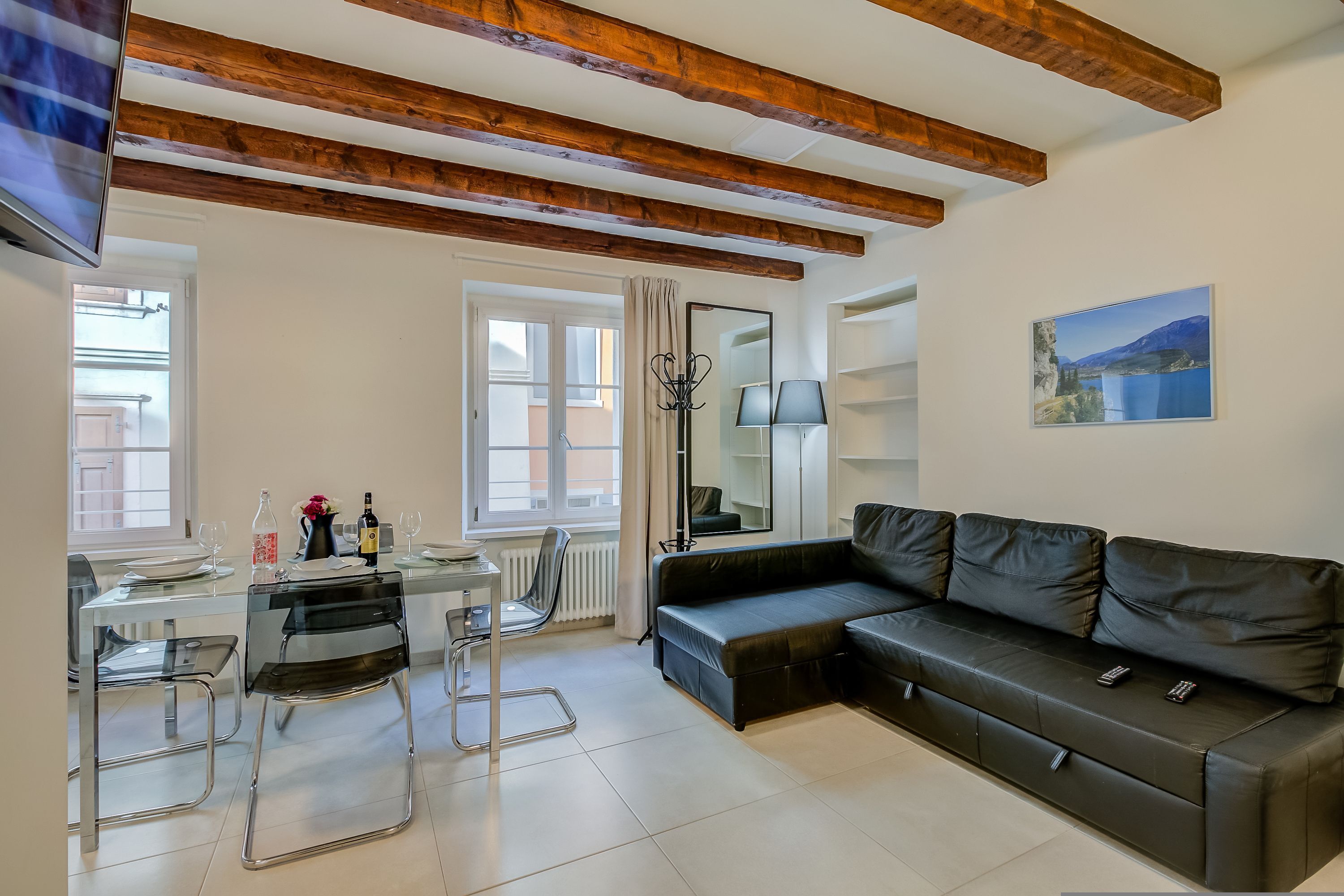 A modern living room with a black leather sofa, a glass dining table with chairs, a TV mounted on the wall, and a window with curtains.