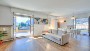 A spacious living room with a sofa, coffee table, and a dining area with a table and chairs. Large windows offer a view of the outdoors. - Antares in Riva del Garda (Riva del Garda)