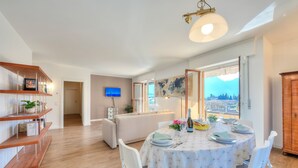 A dining area with a round table set for four, white chairs, a television on the wall, and a view of the city through large windows. - Antares in Riva del Garda (Riva del Garda)
