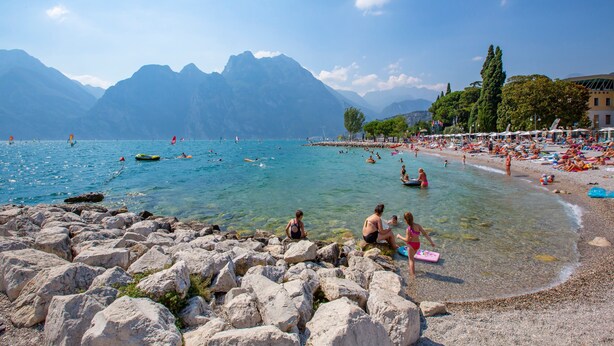 Une plage de galets avec une eau cristalline, des montagnes en arrière-plan et des gens qui profitent de la plage.