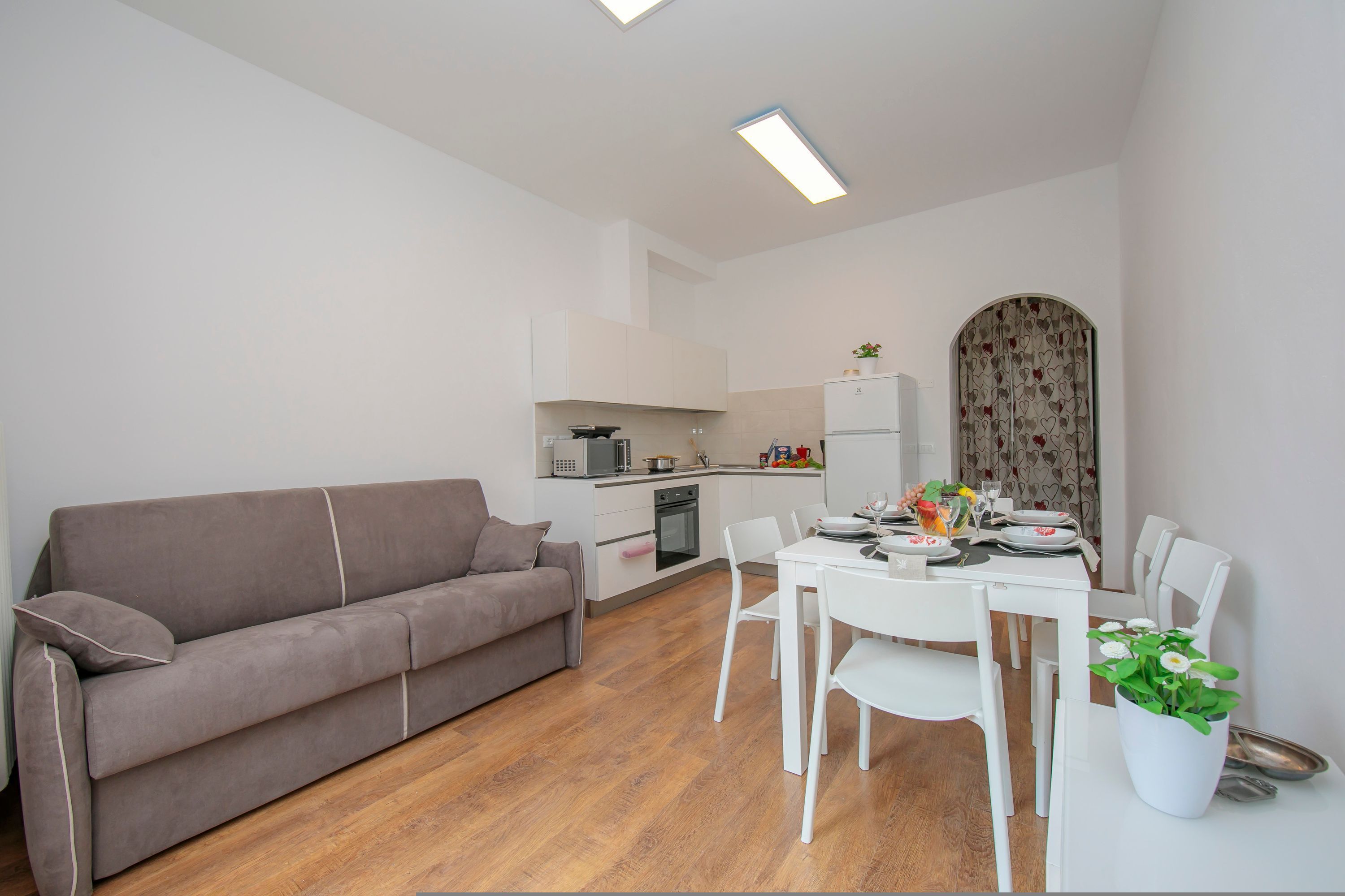 A modern kitchen with white cabinets, a microwave, and a stove. There is a dining area with a white table and chairs, and a gray sofa.