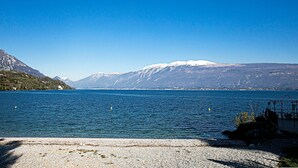 A lake with a rocky shore, clear blue water, and snow-capped mountains in the background. - Borgo Degli Ulivi in Toscolano Maderno (Toscolano Maderno)
