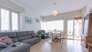A living room with a grey sofa, a dining table with chairs, and a large window with curtains. - Residenza La Terrazza di Dante (Stresa)