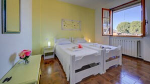 Ein Zimmer mit zwei Betten, einem Nachttisch mit Blumenvase, einer Nachttischlampe und einem Fenster mit Blick auf Bäume.