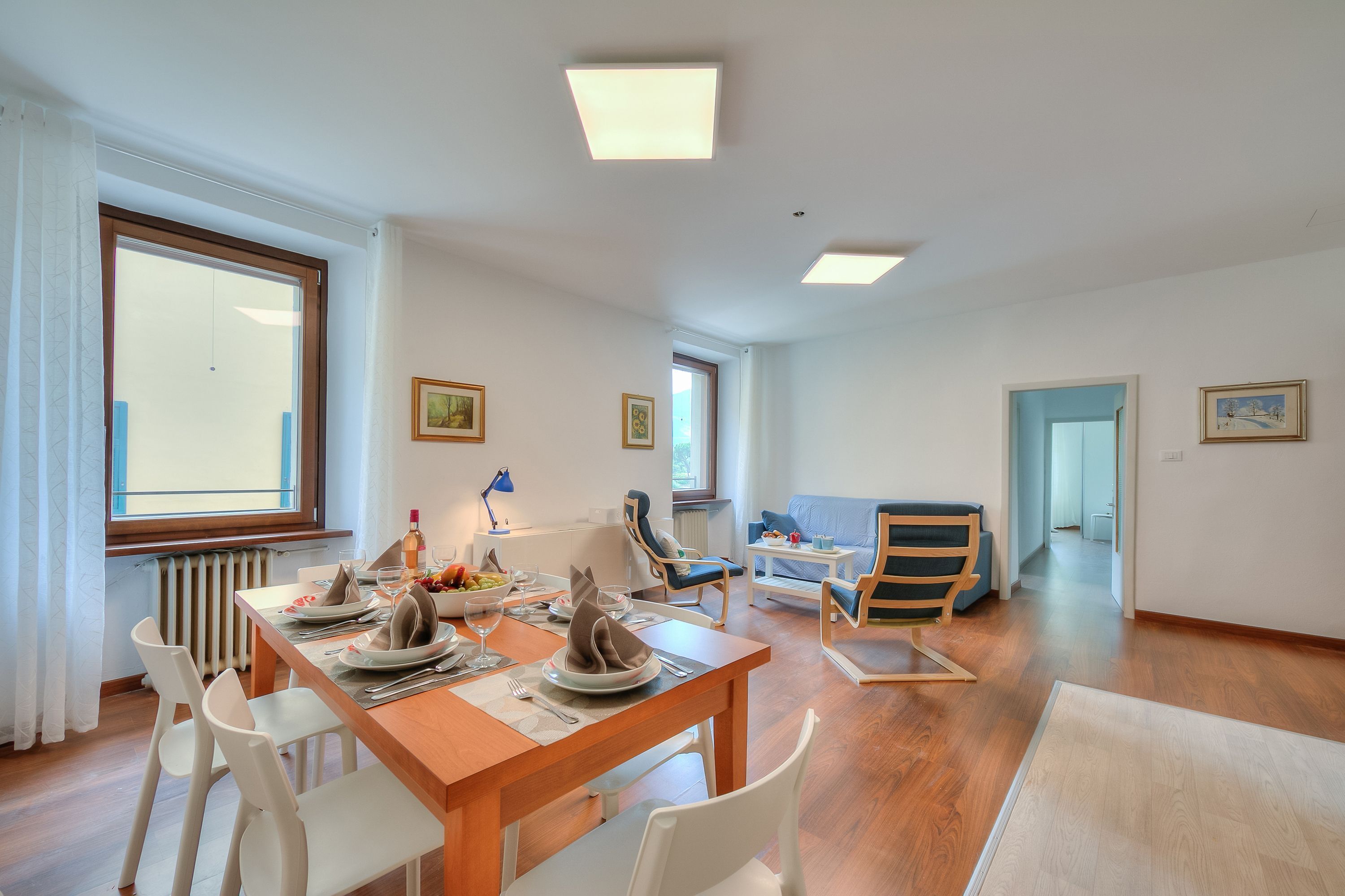 A dining area with a wooden table set for four, white chairs, and a view through a large window.