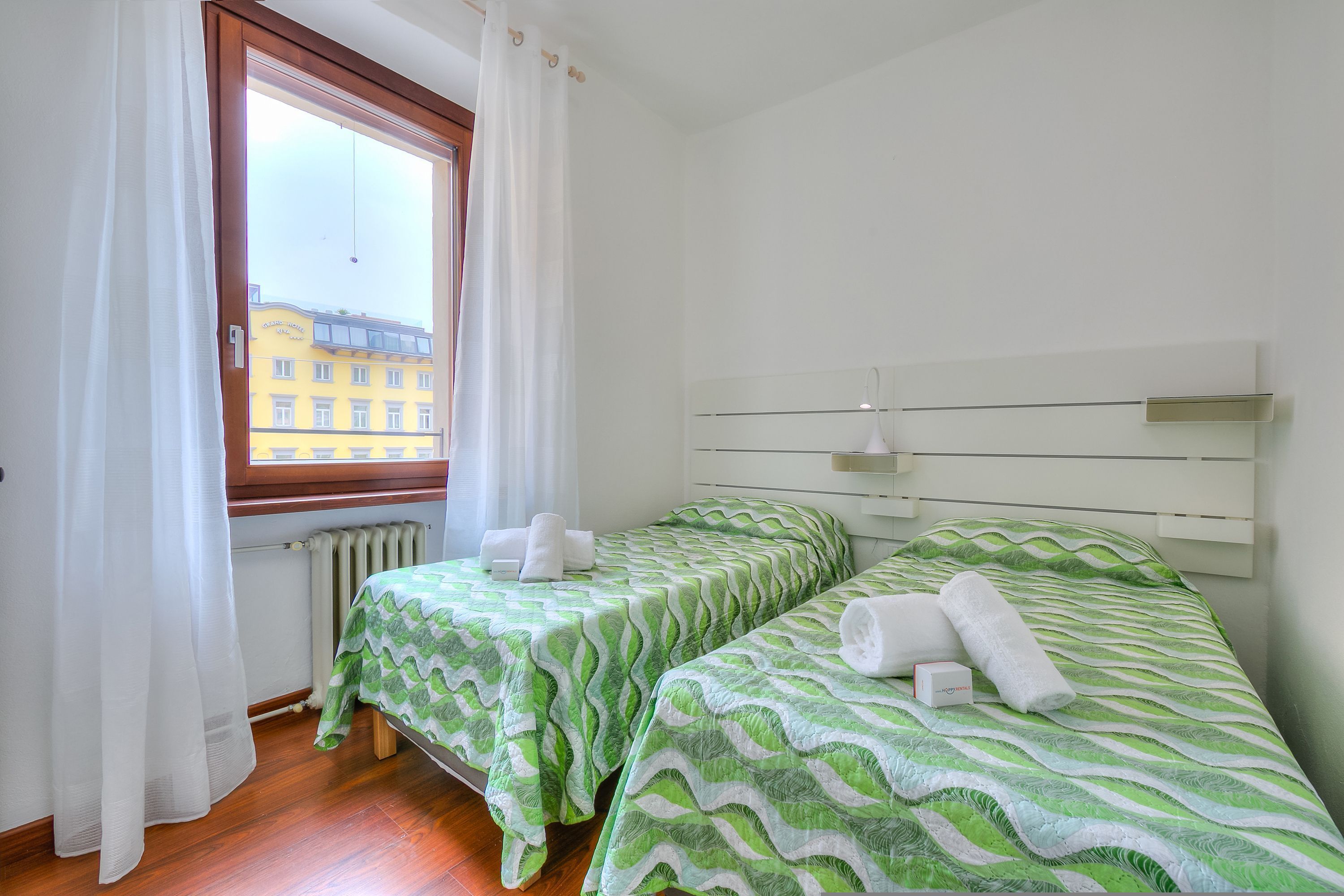 A room with two beds, each with a green patterned bedspread and white towels. There is a window with white curtains, a radiator, and a white headboard with a light fixture.