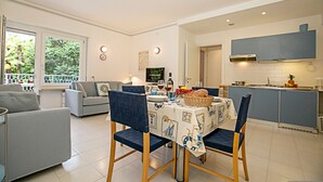 A modern kitchen with a dining area, featuring a table set for a meal, a sofa, and a view of greenery outside. - Riva Lago e Spiaggia Apartment 3 (Riva del Garda)