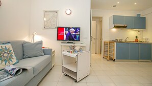 A modern living room with a grey sofa, a television, a small white cart, and a kitchen area with blue cabinets. - Riva Lago e Spiaggia Apartment 3 (Riva del Garda)