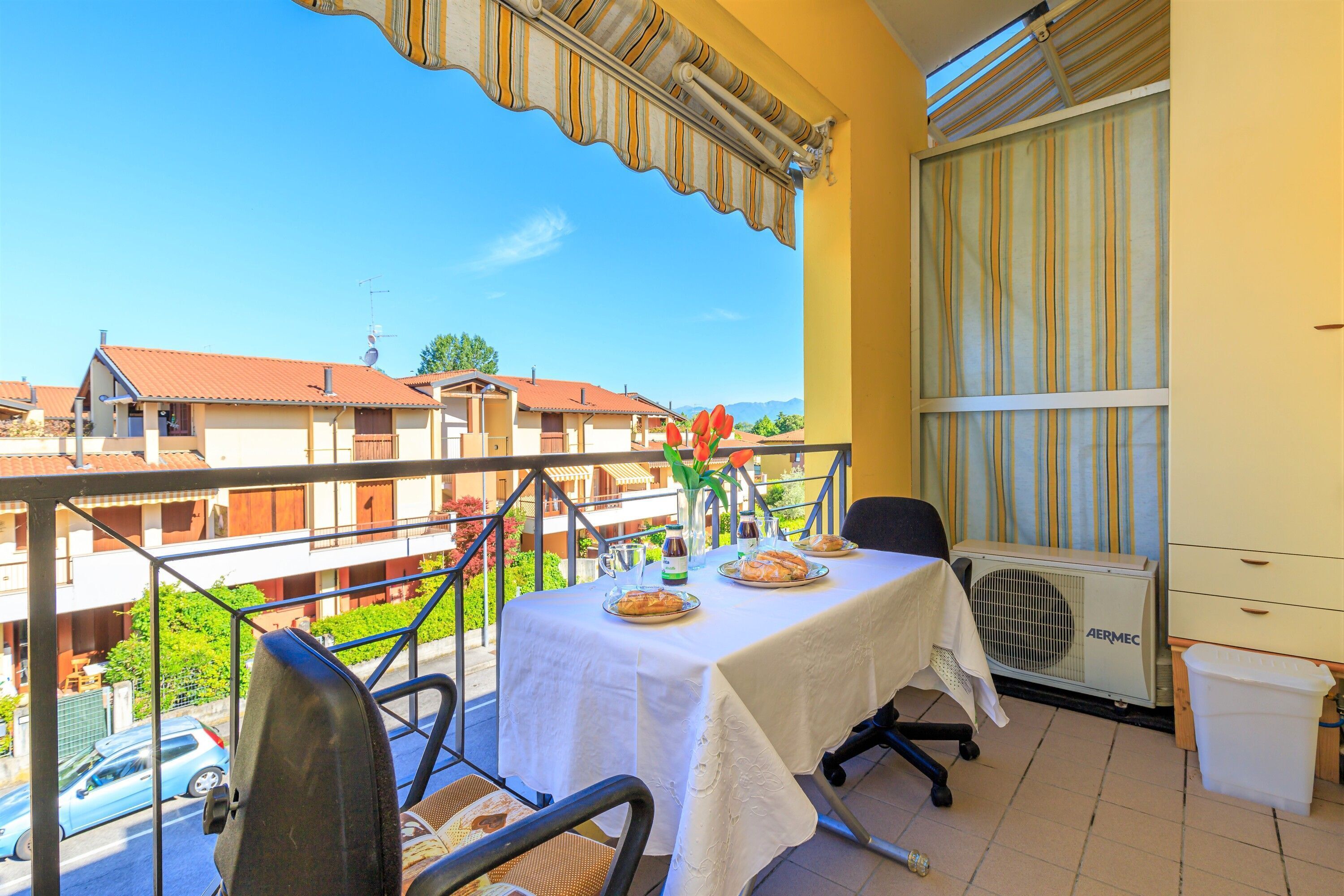 A balcony with a table set for two, chairs, a small table with a vase, and an air conditioning unit.