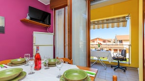 Een eethoek met een tafel gedekt voor vier personen, een televisie aan een roze muur, en een balkon met uitzicht op gebouwen.
