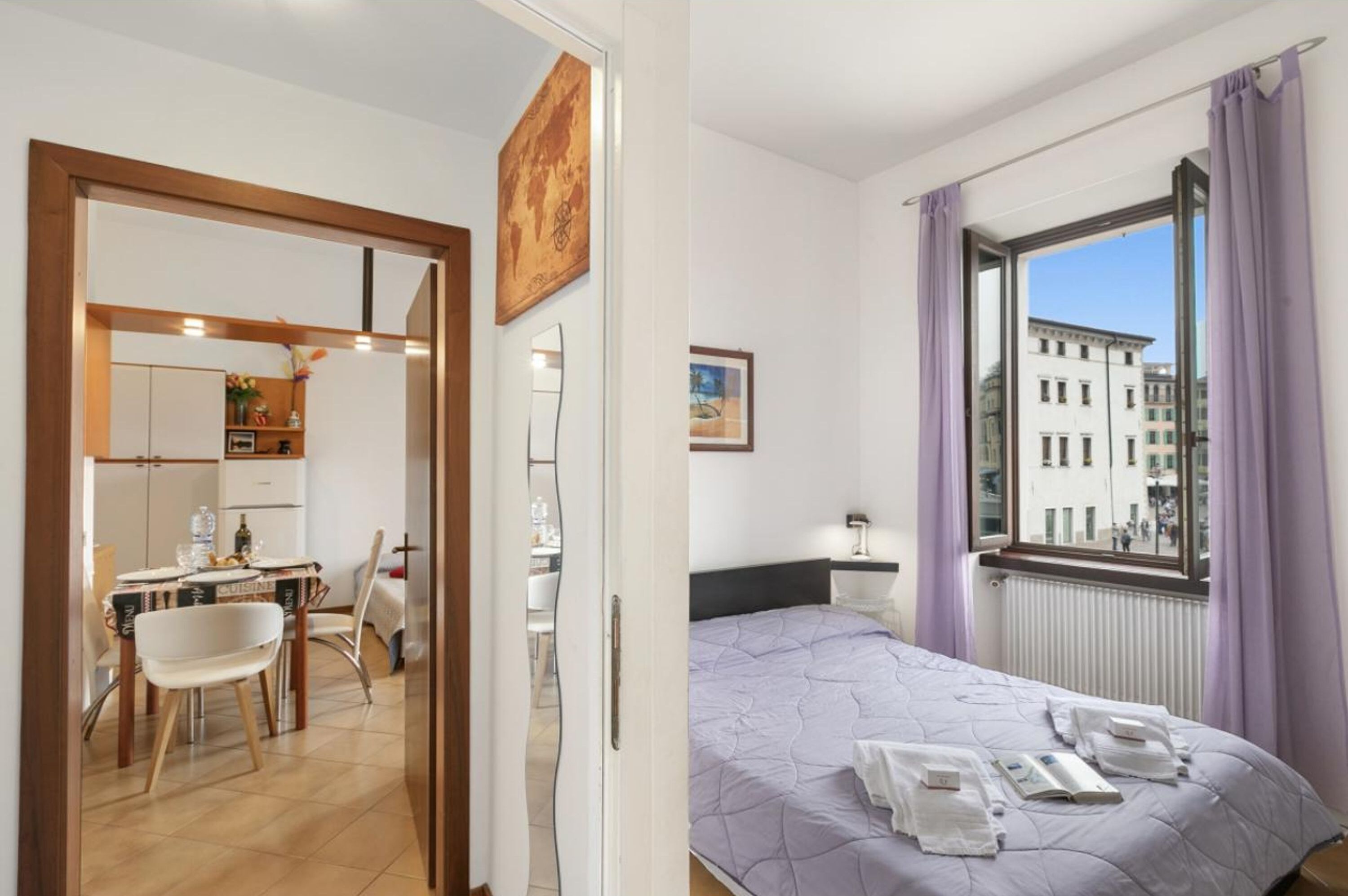 Une chambre d’hôtel avec un lit, une fenêtre avec des rideaux, une petite table avec une chaise, et une vue sur les bâtiments à l’extérieur.