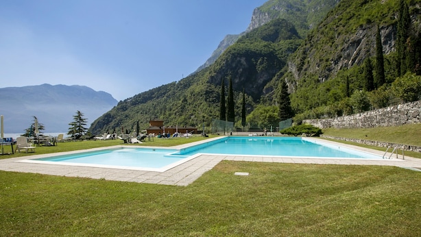 A rectangular swimming pool with a stone deck, surrounded by a grassy area and a stone wall, with mountains in the background. - Lake View Dom-us Apartment (Riva del Garda)