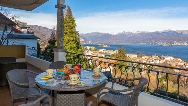 Een balkon met rieten meubels, een tafel met er op een koffiepot, bekers en een schaal fruit.