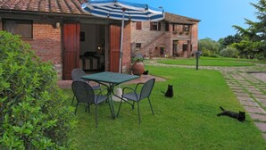 Ein Backsteinhaus mit einer Terrasse, einem Tisch und Stühlen. Eine Katze liegt im Gras.