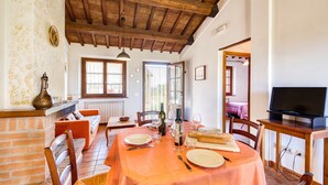 A dining area with a table set for two, a brick fireplace, and a television on a wooden stand. - Myrtle apt With Shared Pool (Castiglione del Lago)
