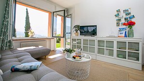 Un soggiorno moderno con un divano grigio, un tavolino rotondo con una torta, una TV su un mobile bianco e vista su un albero attraverso una grande finestra.