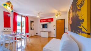 A modern living room with a white sofa, a dining area with a table set for four, a kitchen area with a TV, and a red sign reading "Tavola calda". - Claz House in Riva del Garda (Riva del Garda)