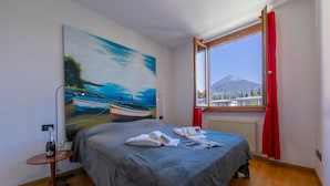 A hotel room with a bed, a painting of boats, a window with a mountain view, and a table with a bottle and glasses. - Claz House in Riva del Garda (Riva del Garda)