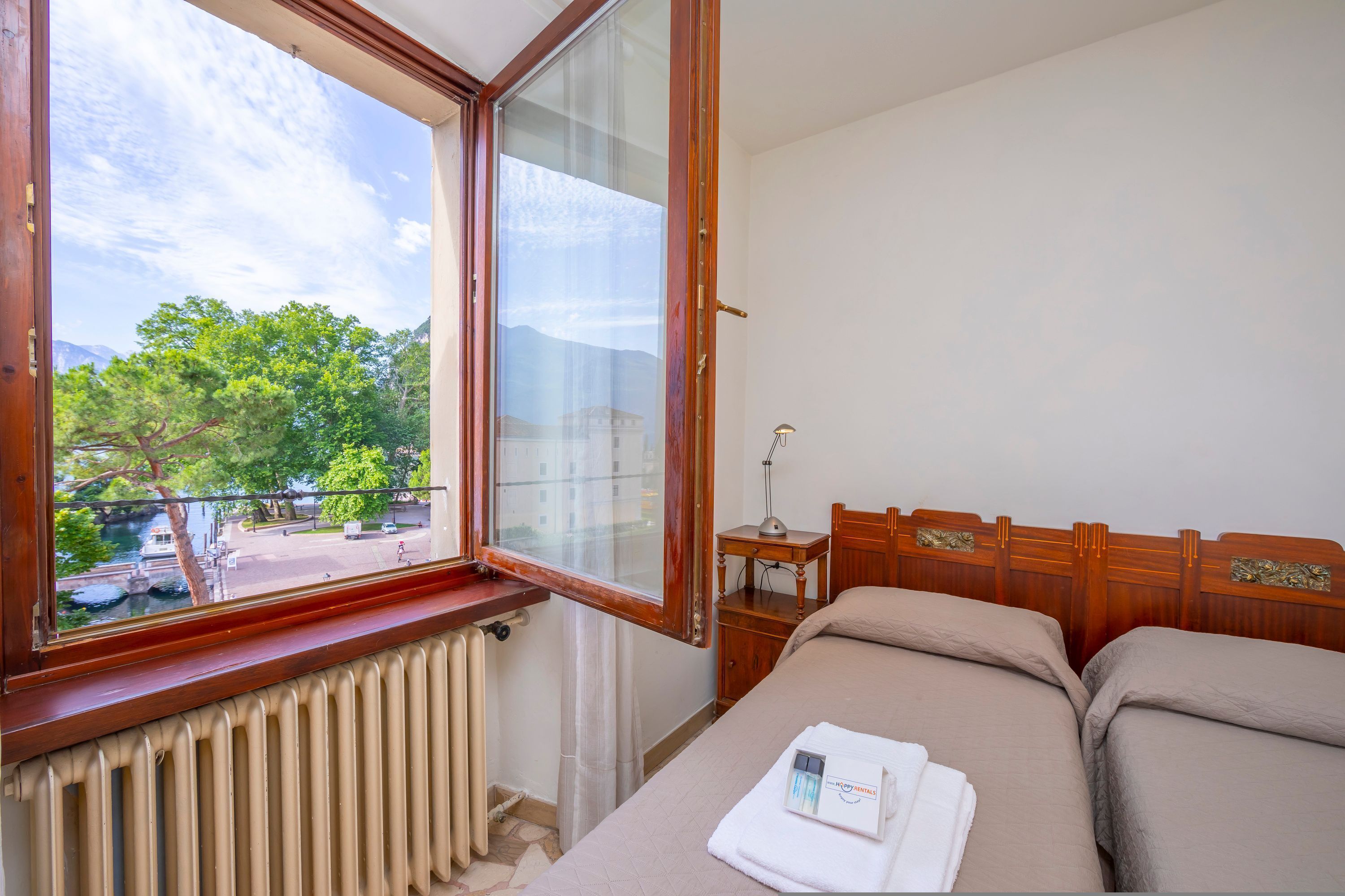 A hotel room with a large window, a radiator, a bedside table with a lamp, and two beds.