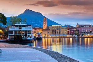 A waterfront hotel with a clock tower, a boat docked nearby, and a mountain in the background. - Residenza Rocca del Lago 3 (Riva del Garda)
