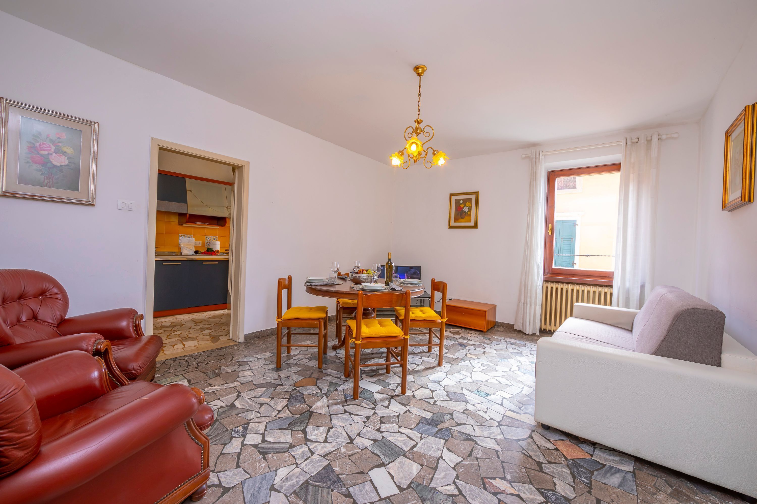 A living room with a stone-tiled floor, a white sofa, a red leather chair, a dining table with chairs, a chandelier, and a kitchen area visible through an open door.