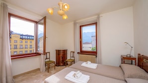Ein Hotelzimmer mit zwei Betten, einer Holzkommode, einem Schreibtisch mit Lampe und großen Fenstern mit Blick auf die Stadt.