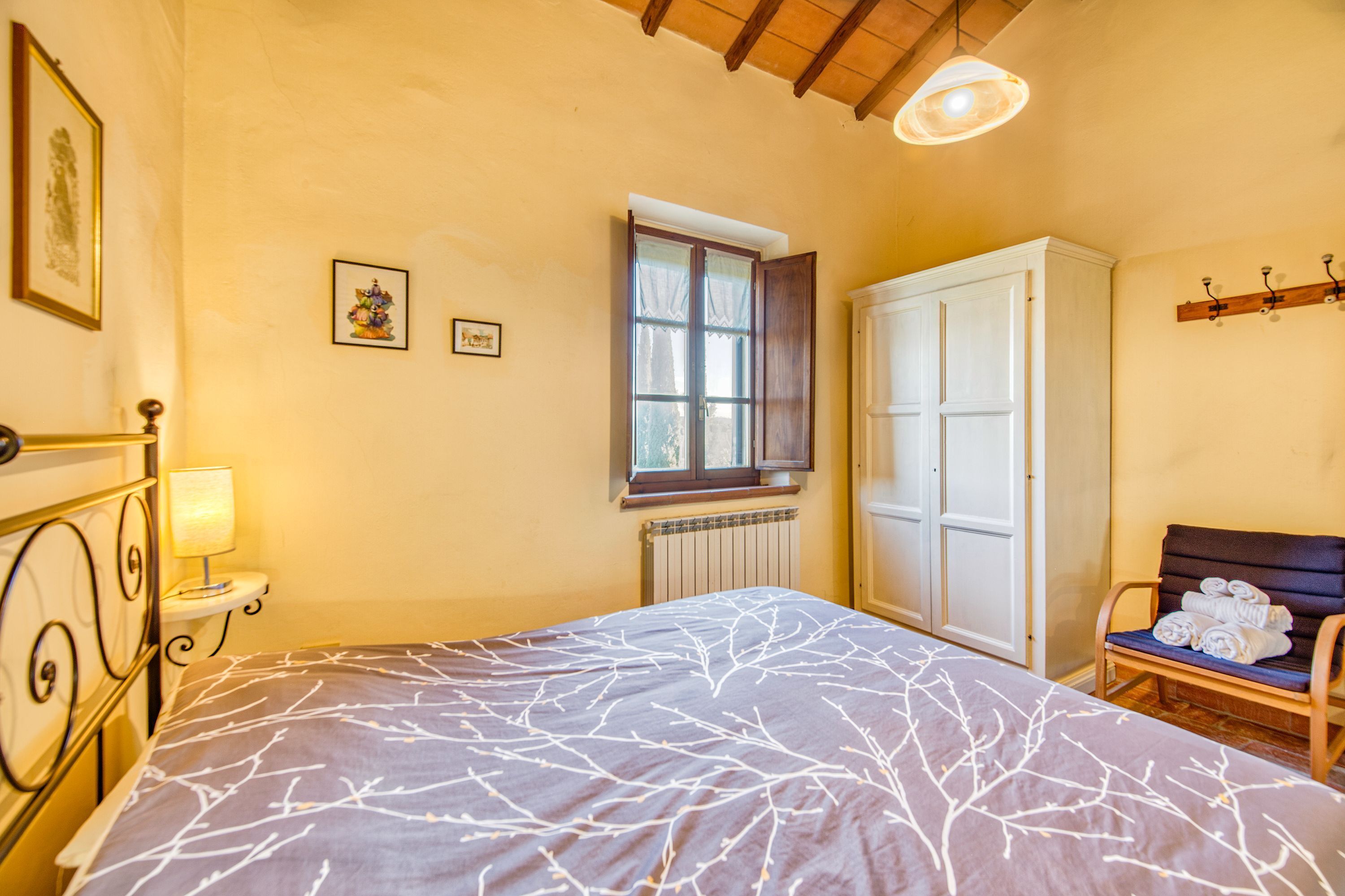 A bedroom with a bed, a radiator, a window, a wardrobe, a chair with towels, and wall-mounted pictures.
