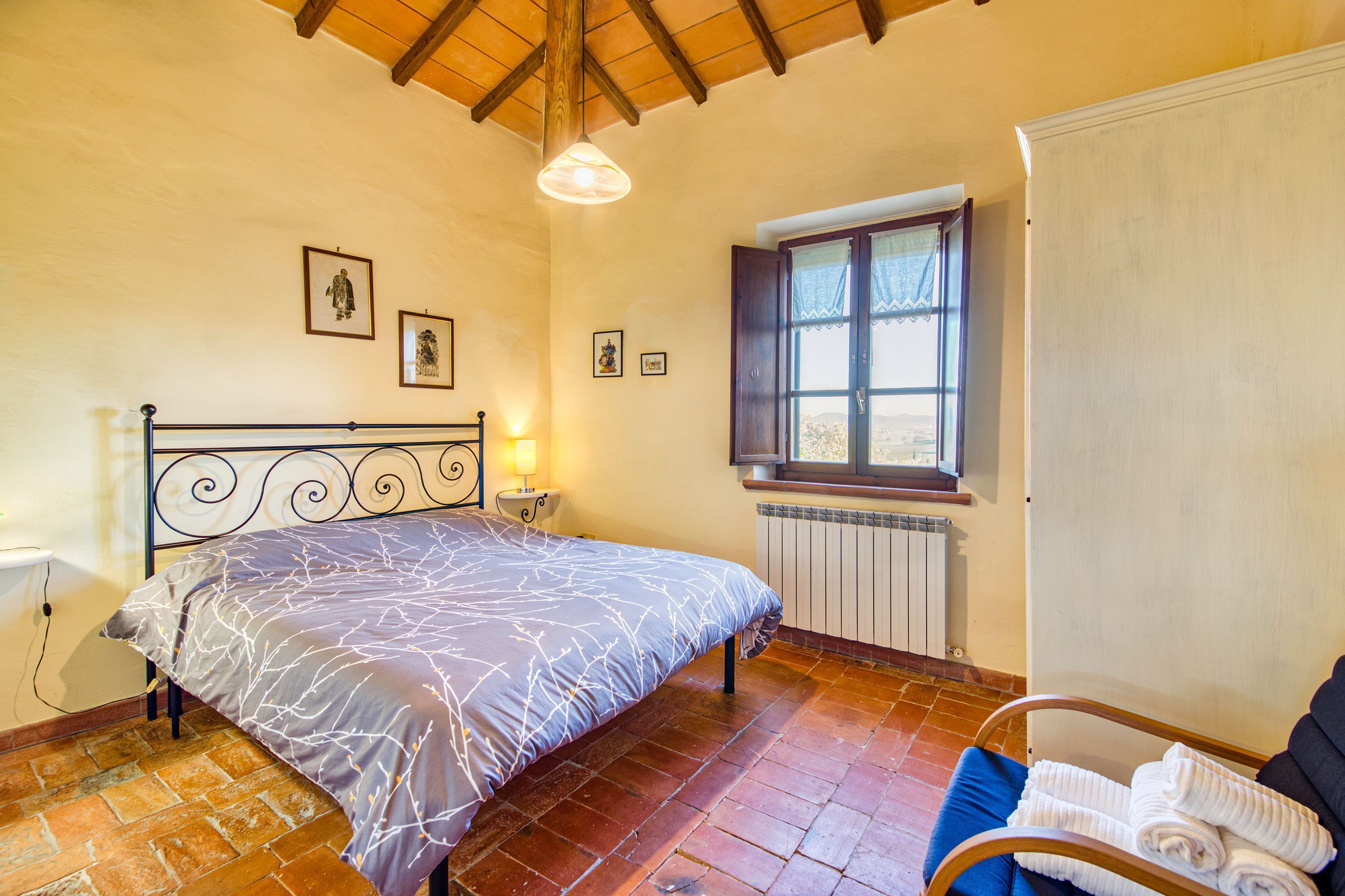 A bedroom with a bed, a window with curtains, a radiator, and a chair with towels.