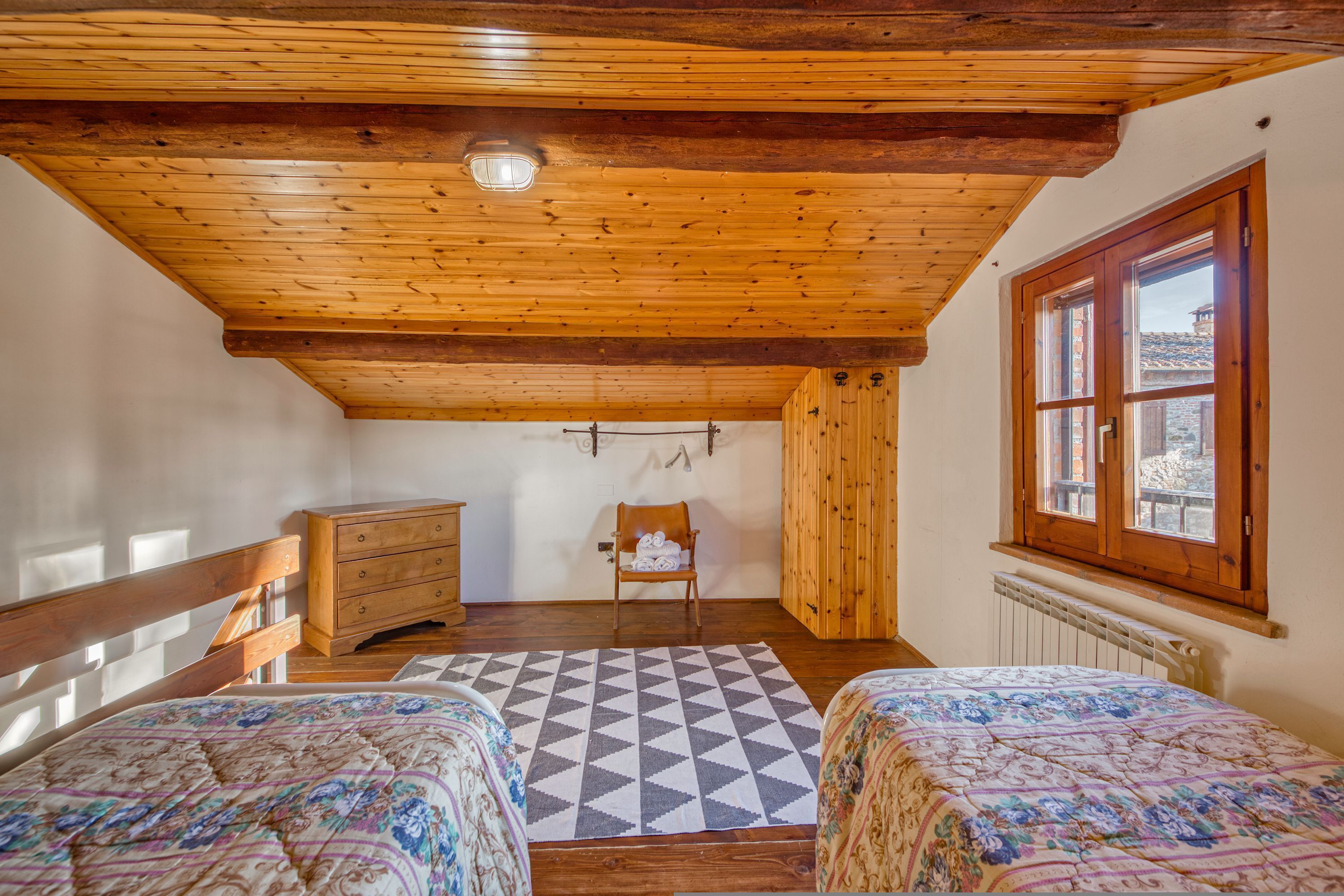 Ein Zimmer mit Holzbalkendecke, zwei Betten mit geblümten Bettdecken, ein Holzkommode, ein Stuhl und ein Fenster mit Blick auf Gebäude.