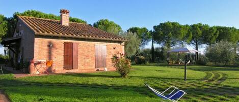 Une maison de campagne en briques, avec un toit en tuiles, un petit jardin avec une pelouse et une chaise pliante bleue.