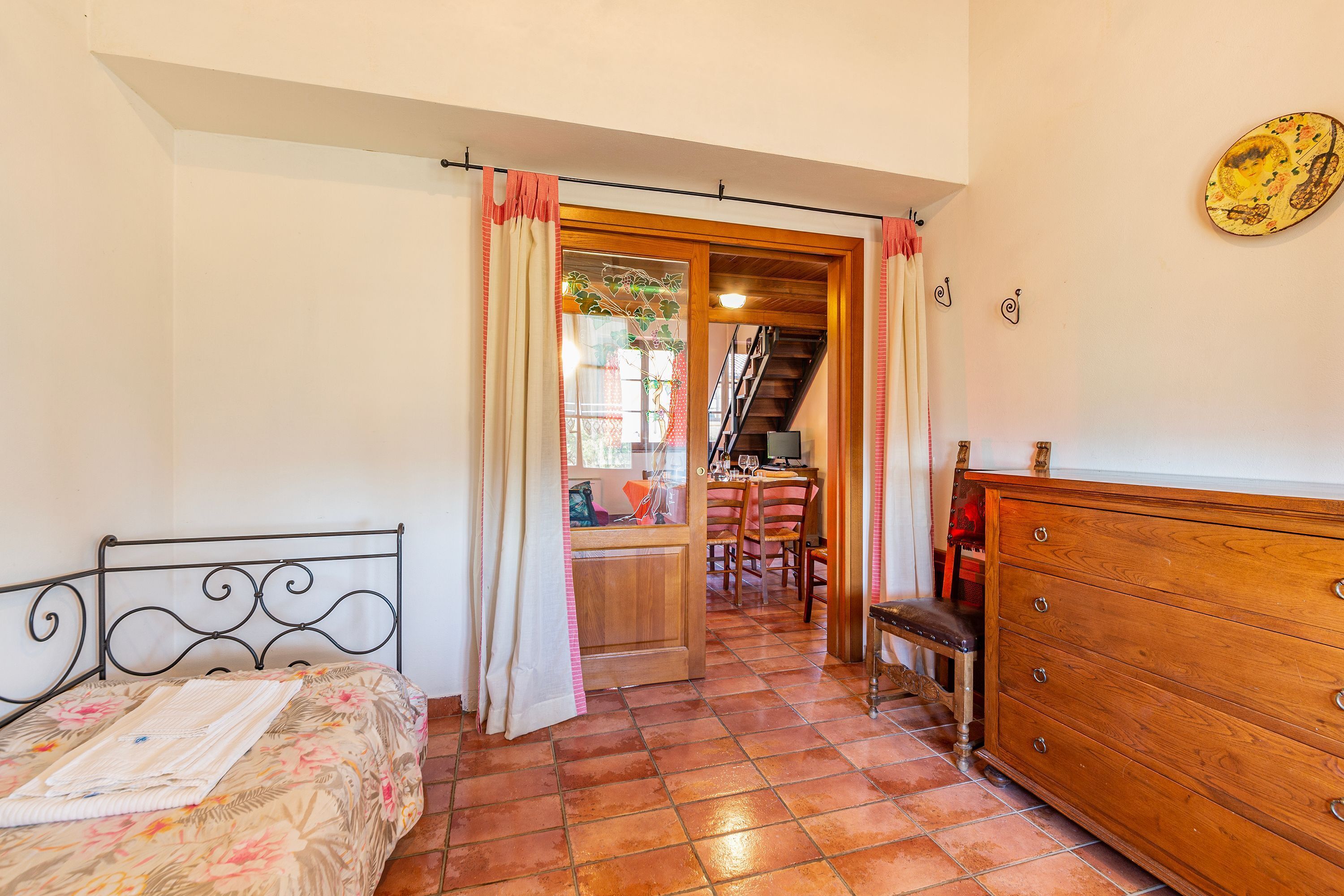 A bedroom with a bed, a dresser, a chair, and a dining table with chairs. There is a staircase leading to another room.
