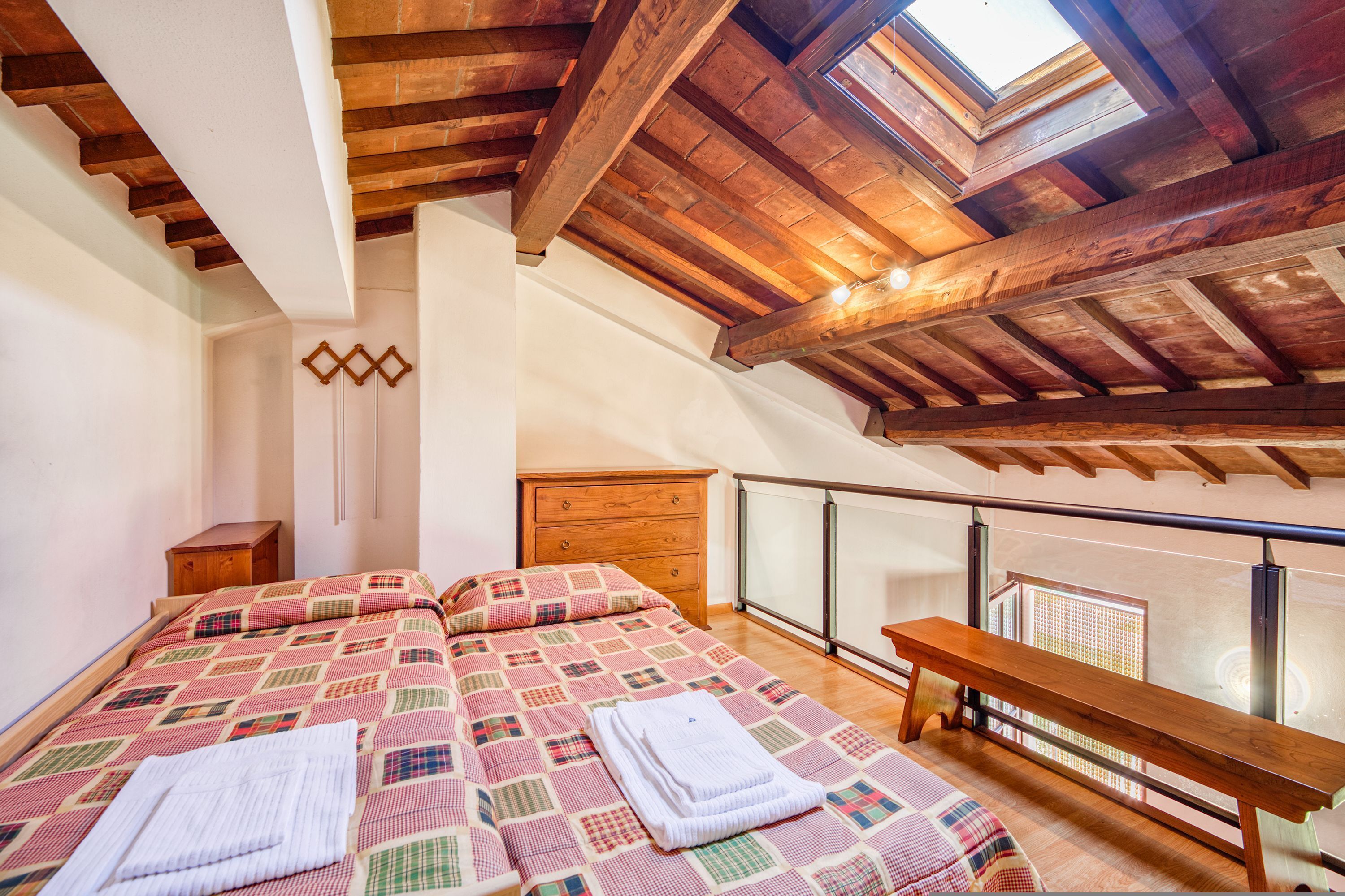 A bedroom with a wooden ceiling, a bed with a checkered bedspread, a bedside table, and a bench by the window.