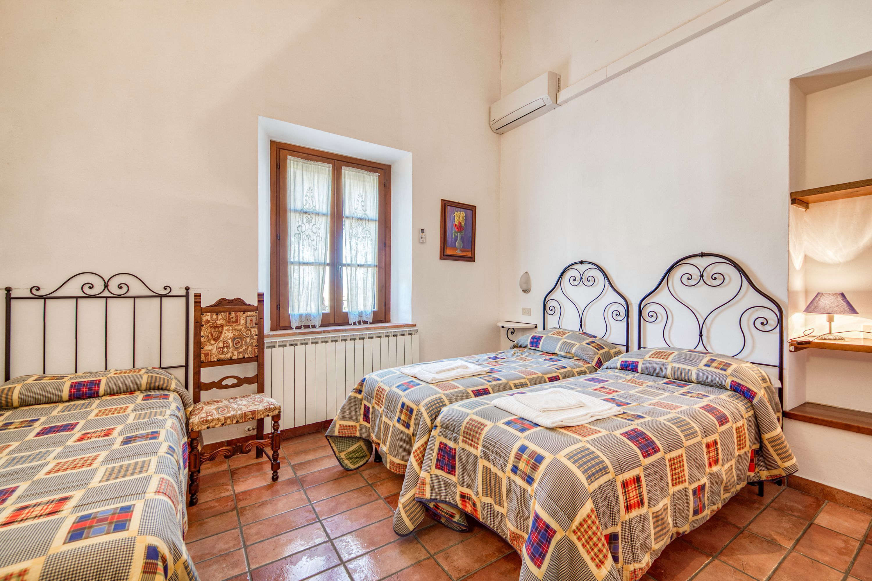 Un dormitorio con dos camas, una silla, una ventana con cortinas y un aire acondicionado de pared.