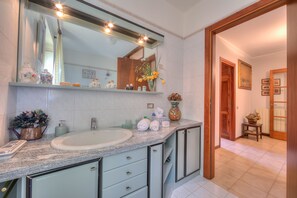 A bathroom with a large mirror, a sink, and a vanity with drawers. There are toiletries on the counter and a vase with flowers. - Casa Elisabetta in Stresa (Stresa)