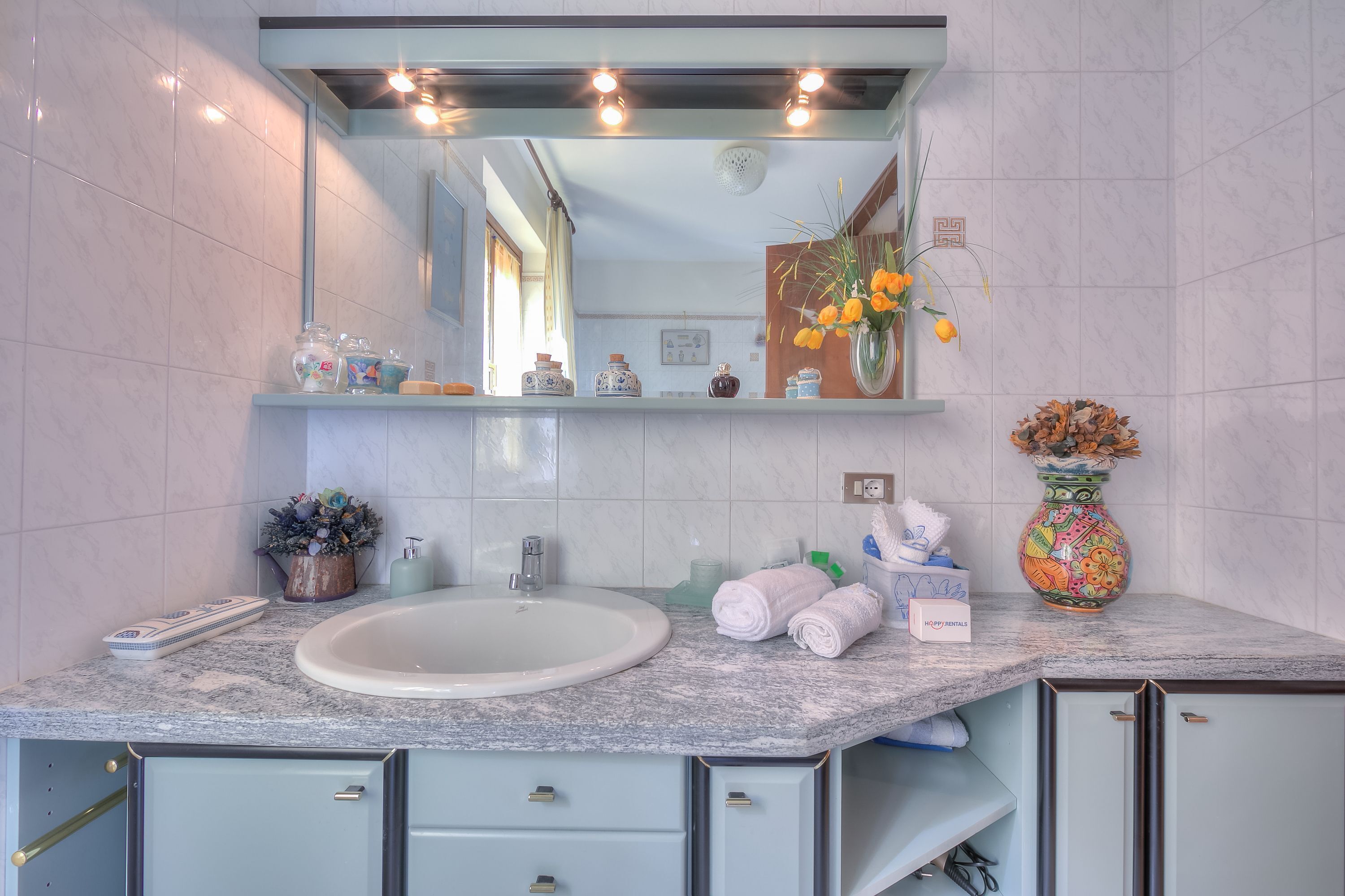 A bathroom with a marble sink, a mirror above it, and a shelf with toiletries and a vase of flowers.