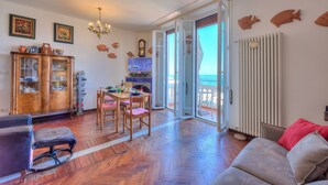 A spacious living room with a wooden floor, a sofa, a dining table with chairs, a cabinet with glass doors, a television, and a view through the glass doors to the outside. - La Vie Enchanting Sea View (Alassio)