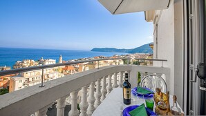 A balcony with a table set for dining, overlooking a coastal cityscape with buildings and the sea. - La Vie Enchanting Sea View (Alassio)