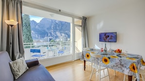 A room with a balcony, a dining table with a sunflower tablecloth, a sofa, and a TV on the wall. - Il Piccolo Nido in Riva del Garda (Riva del Garda)