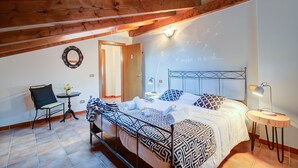 A bedroom with a bed, a chair, a small table, and a wall with a light fixture and a wall art piece. - Villa Oasi With Private Pool (Sirmione)