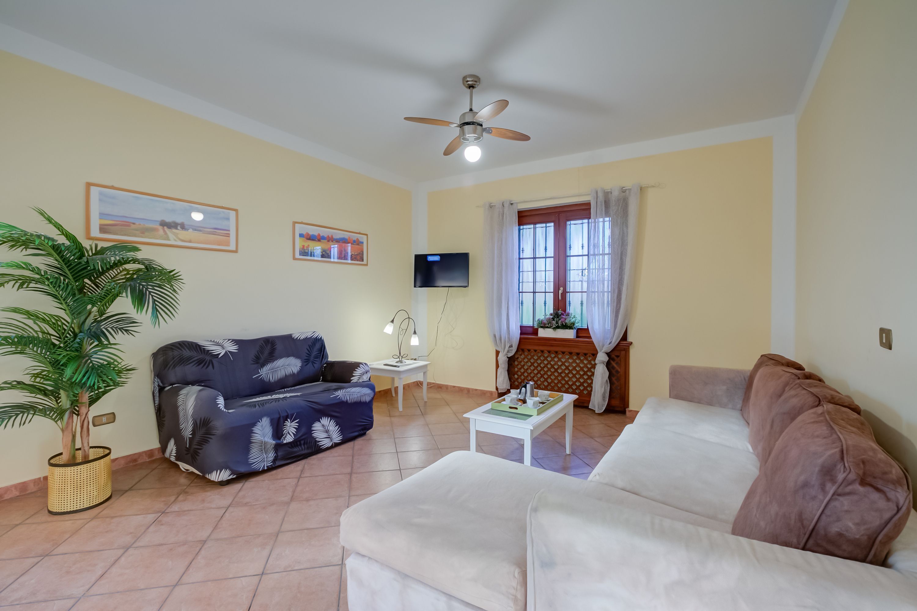 Un salon avec un ventilateur de plafond, deux canapés, une table basse, un téléviseur et une fenêtre avec des rideaux.