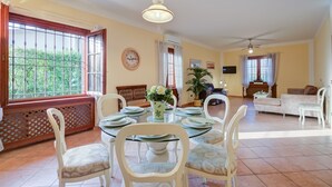 A dining area with a round table, white chairs, and a vase of flowers. There is a sofa and a television in the background. - Villa Oasi With Private Pool (Sirmione)