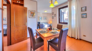 A dining area with a wooden table set for four, surrounded by chairs. There is a window with curtains, a radiator, and framed pictures on the wall.