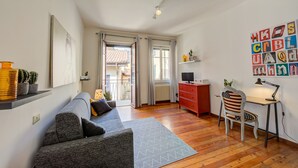 Un salon moderne avec un canapé gris, un plancher en bois, un bureau avec une chaise et une vue sur un espace extérieur, visible à travers une porte.