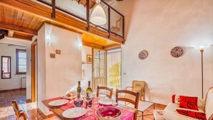 Ein Essbereich mit einem Holztisch, gedeckt für vier Personen, umgeben von Stühlen. Darüber befindet sich ein Balkon, und der Raum hat eine rustikale Einrichtung mit freiliegenden Holzbalken und einem Fliesenboden.