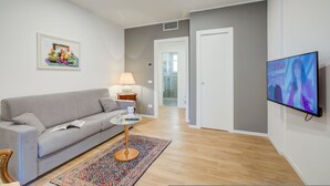 Una sala de estar moderna con un sofá gris, una mesita redonda pequeña, una lámpara, una alfombra colorida y un televisor en la pared.