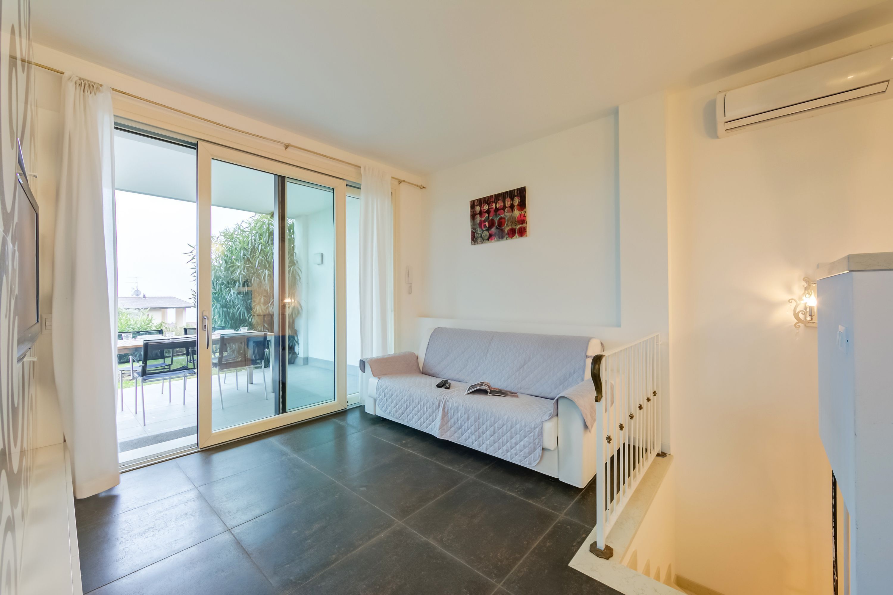 A modern living room with a white sofa, a glass door leading to an outdoor area, and a wall-mounted air conditioning unit.