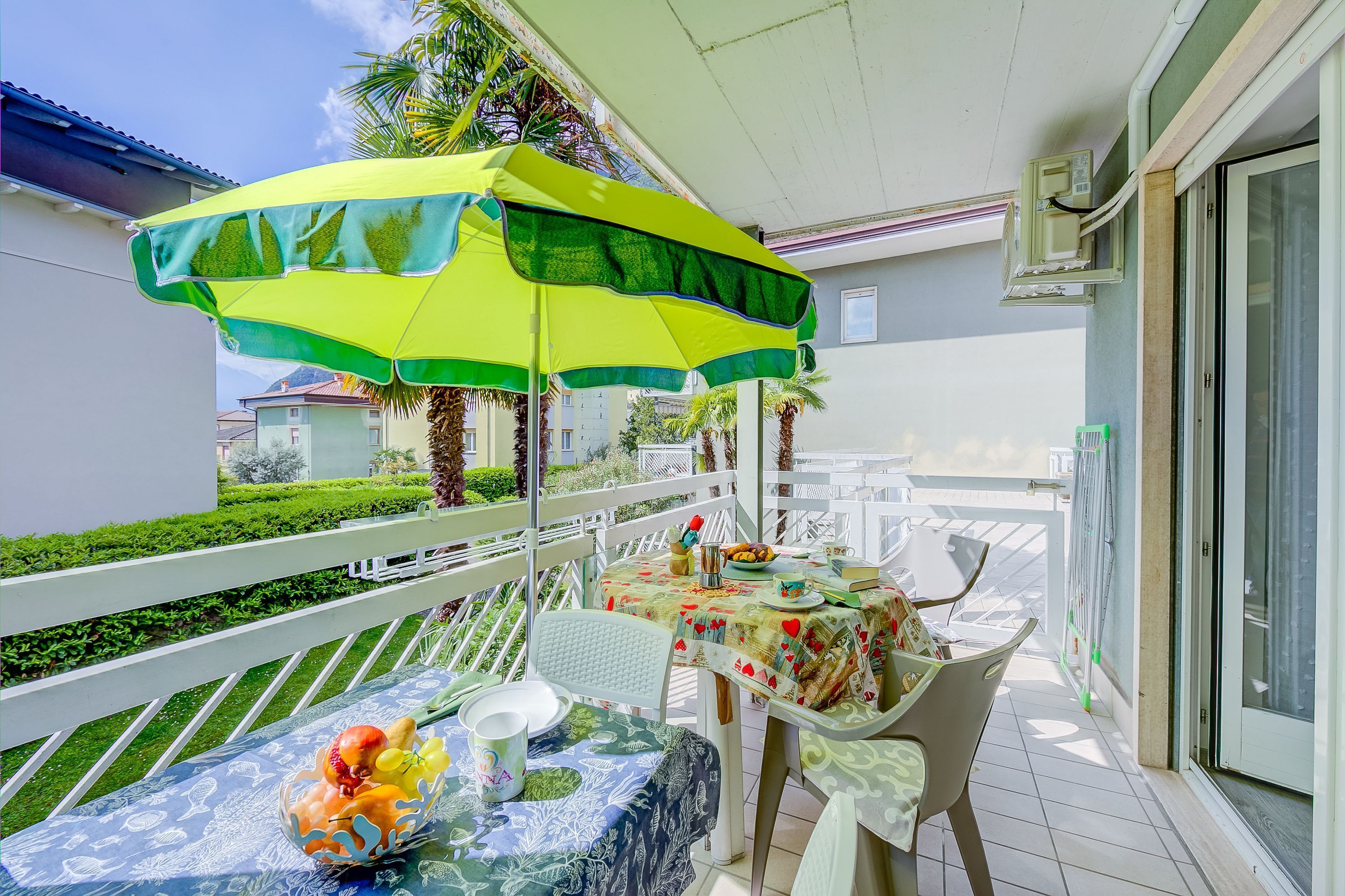 A balcony with a table set for two, covered by a yellow and green umbrella, and a white railing.