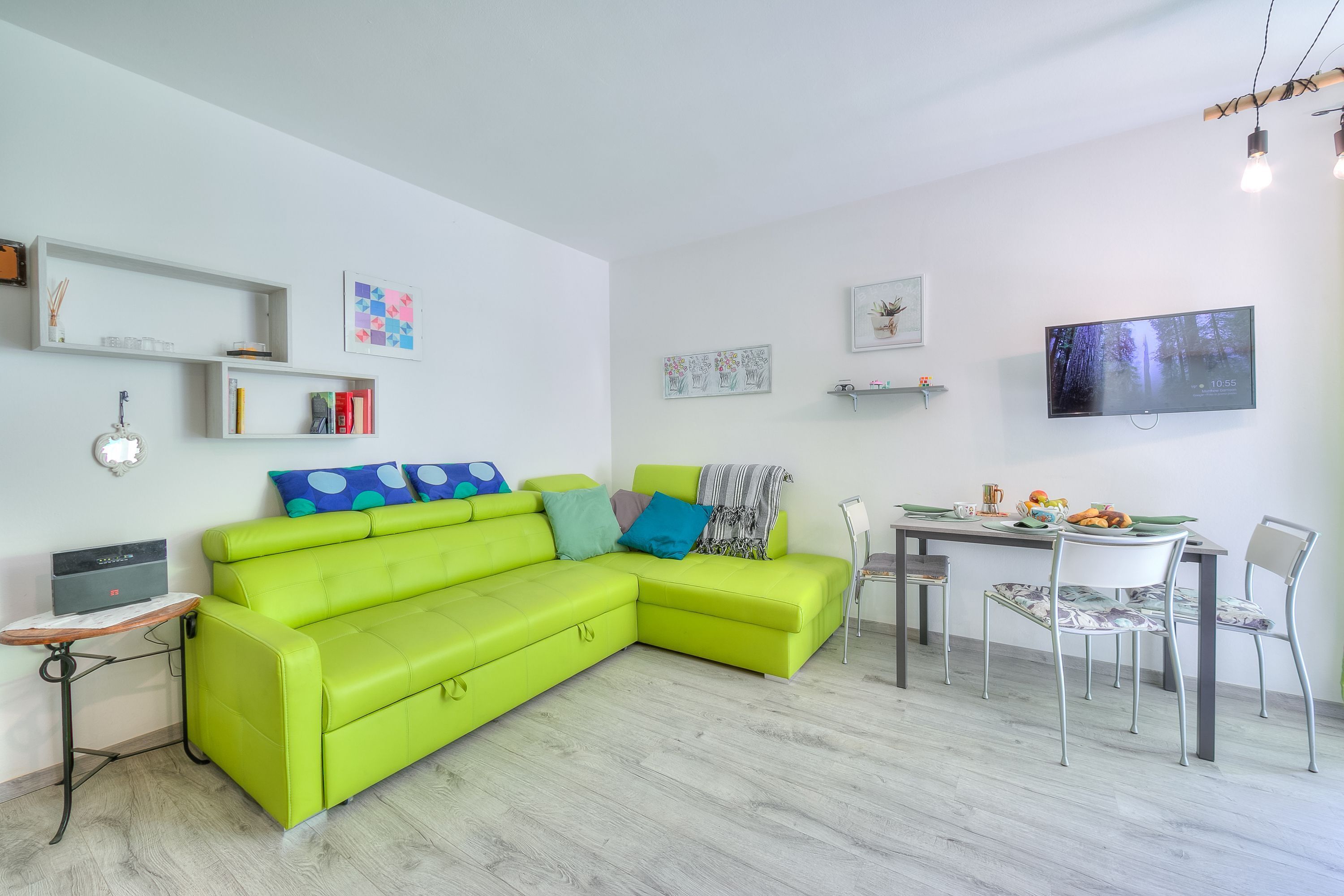 Een lichte woonkamer met een groene bank, een eettafel met stoelen en een flatscreen televisie aan de muur.